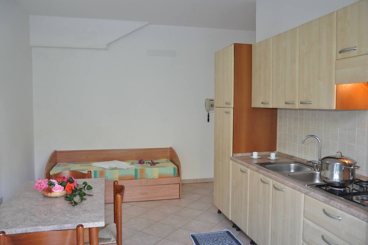 Eine kompakte Küche mit Holzausstattung, Spüle und Herd. Ein kleiner Esstisch mit Stühlen und eine Bank mit Kissen sind vorhanden. Ein Telefon ist an der Wand befestigt.