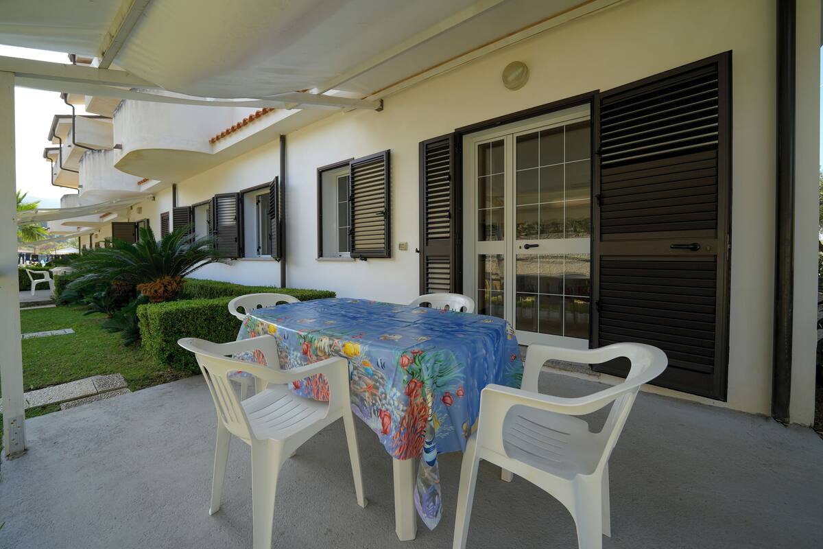 Eine Terrasse im Freien mit Tisch und Stühlen, einem überdachten Bereich und einem Gebäude mit Fensterläden.