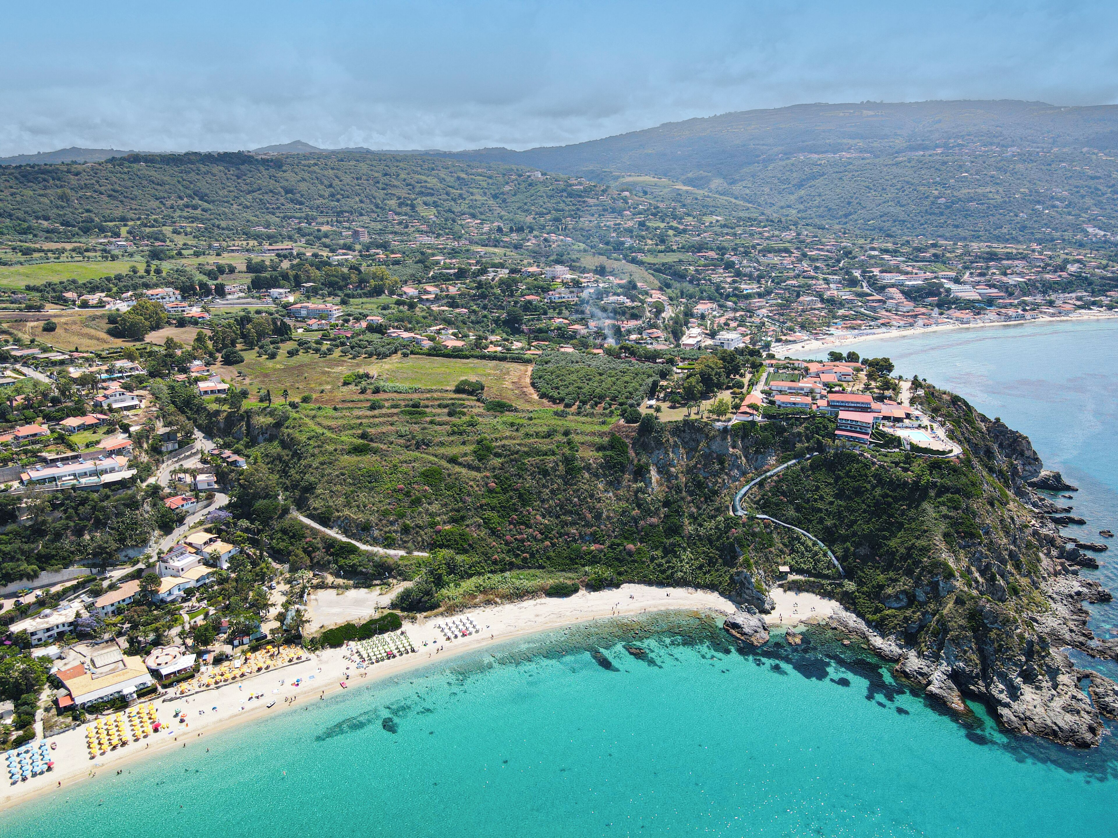 Vista aèria d'una població costanera amb una platja, aigües turqueses cristal·lines i una combinació d'edificis residencials i comercials.