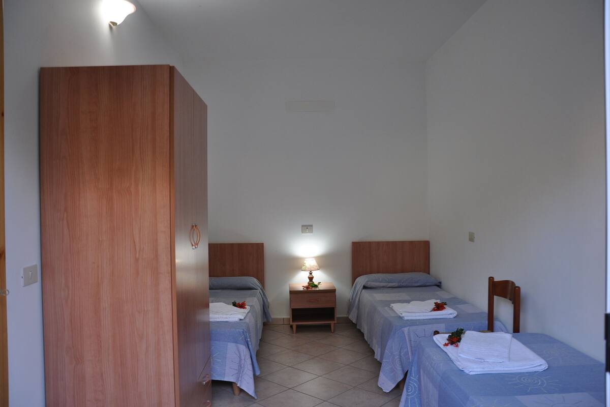 Ein kleines, gut beleuchtetes Zimmer mit zwei Einzelbetten, einem Holzschrank, einem Nachttisch mit Lampe und einem Stuhl.