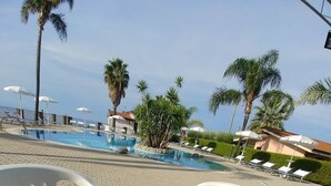 A table with two cups of coffee and a croissant, overlooking a pool area with lounge chairs and palm trees. - Hotel Residence Rosy (Ricadi)