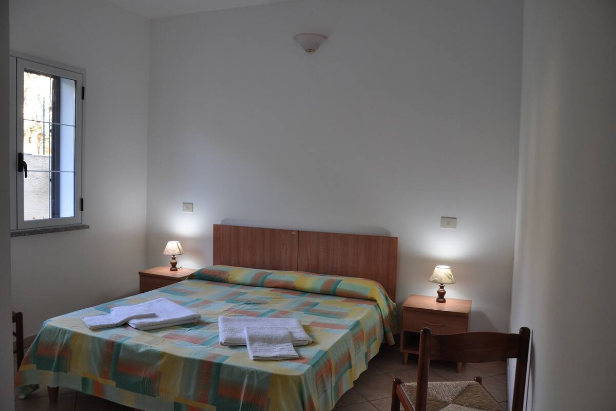 Ein Schlafzimmer mit einem Bett, zwei Nachttischlampen, einem Holznachttisch, einem Sessel und einem Fenster.