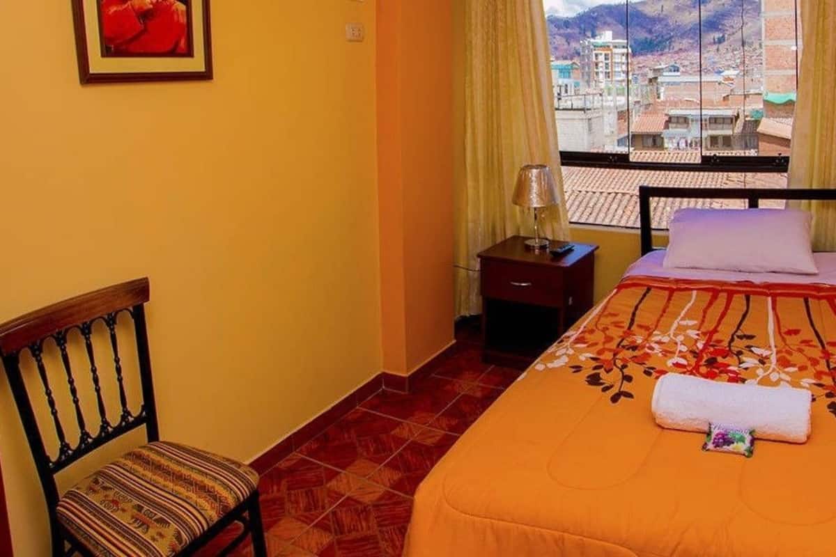A small hotel room with a bed, a chair, a nightstand, and a window with a view of buildings.