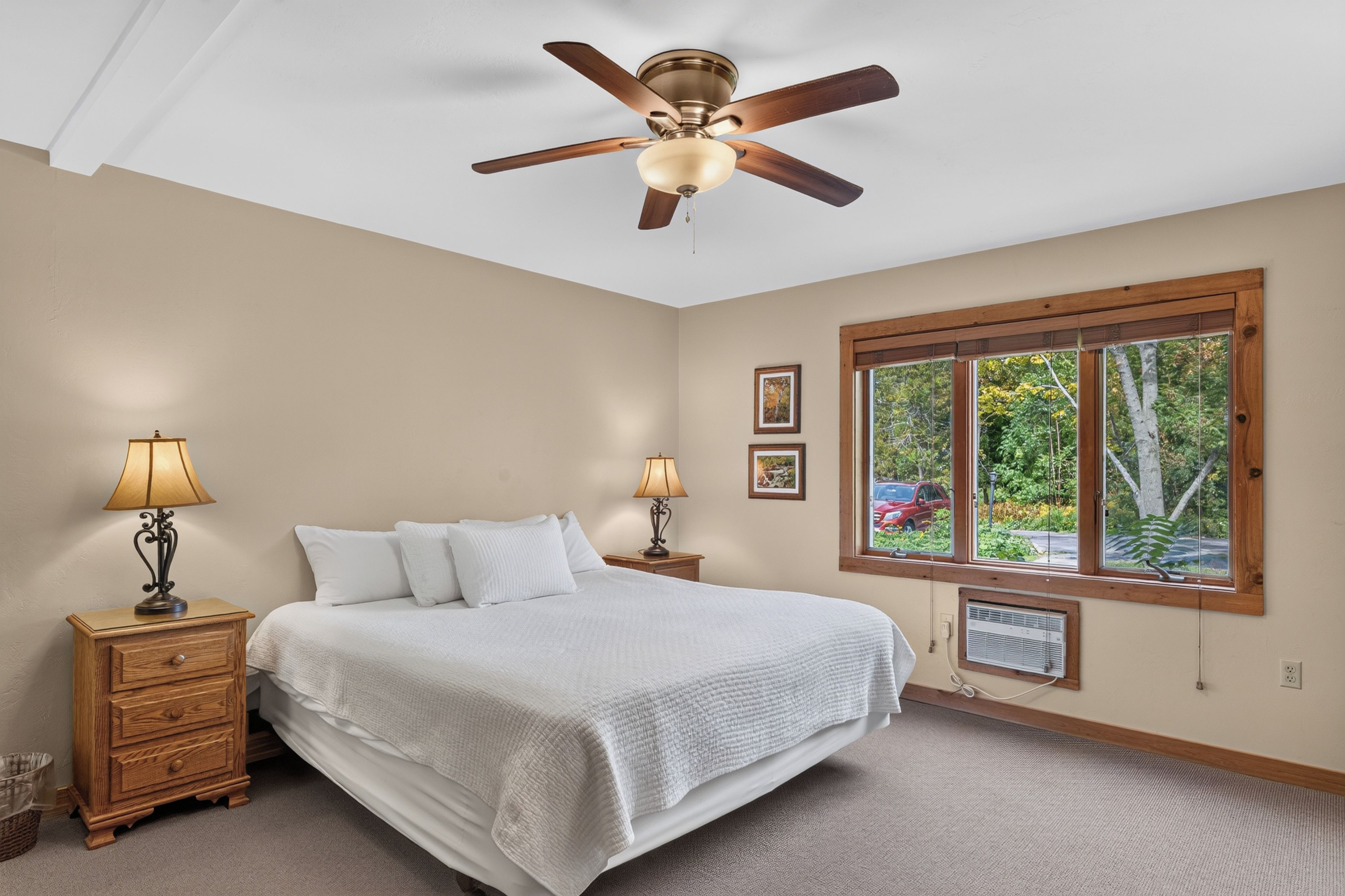 A bedroom with a bed, two bedside tables with lamps, a ceiling fan, a window with a view of trees, and a wall-mounted air conditioner.