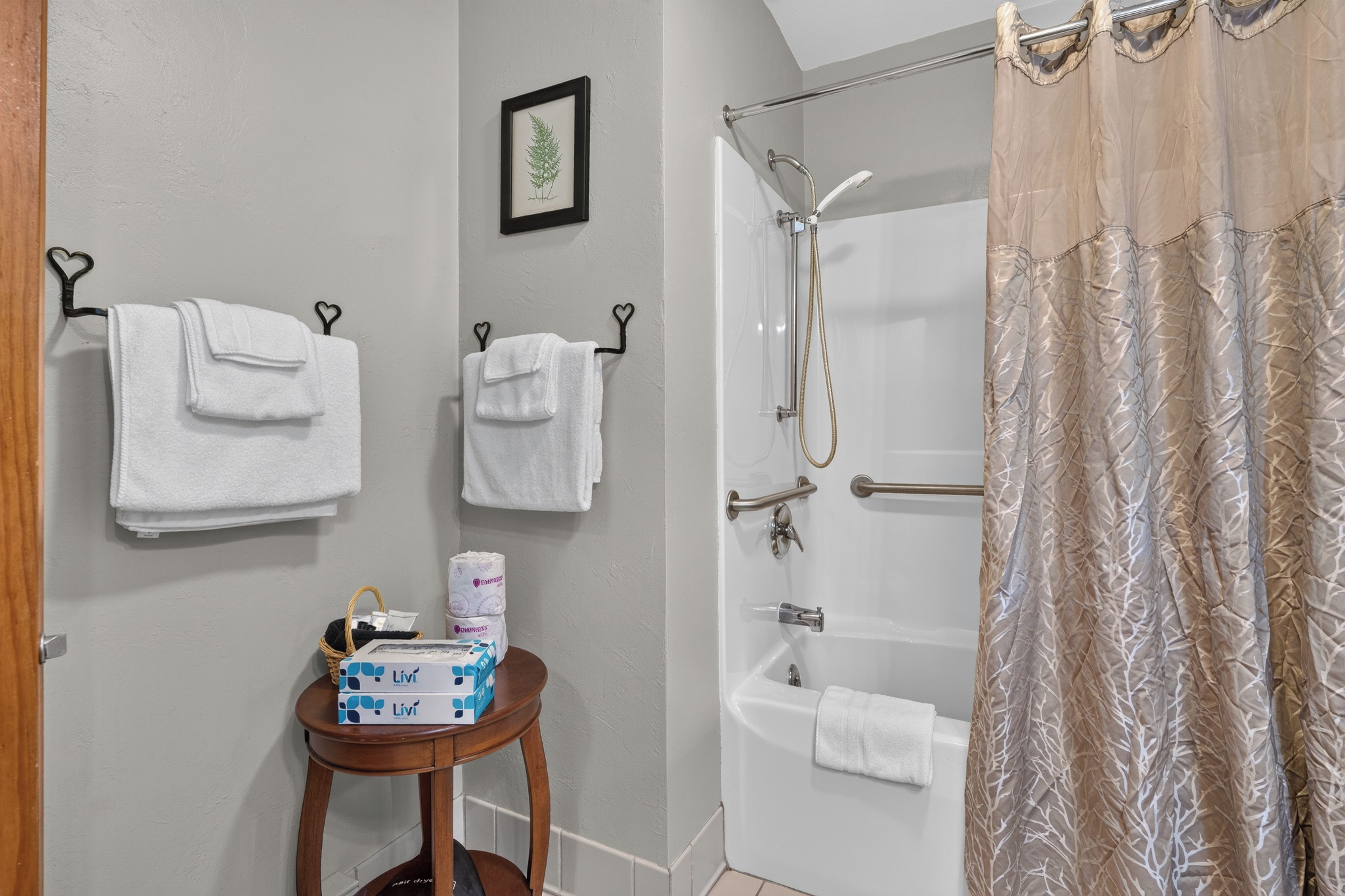 A bathroom with two white towels hanging on hooks, a framed botanical illustration on the wall, a small wooden table with toiletries, and a shower with a beige curtain.
