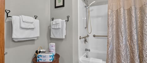 A bathroom with two white towels hanging on hooks, a framed botanical illustration on the wall, a small wooden table with toiletries, and a shower with a beige curtain.