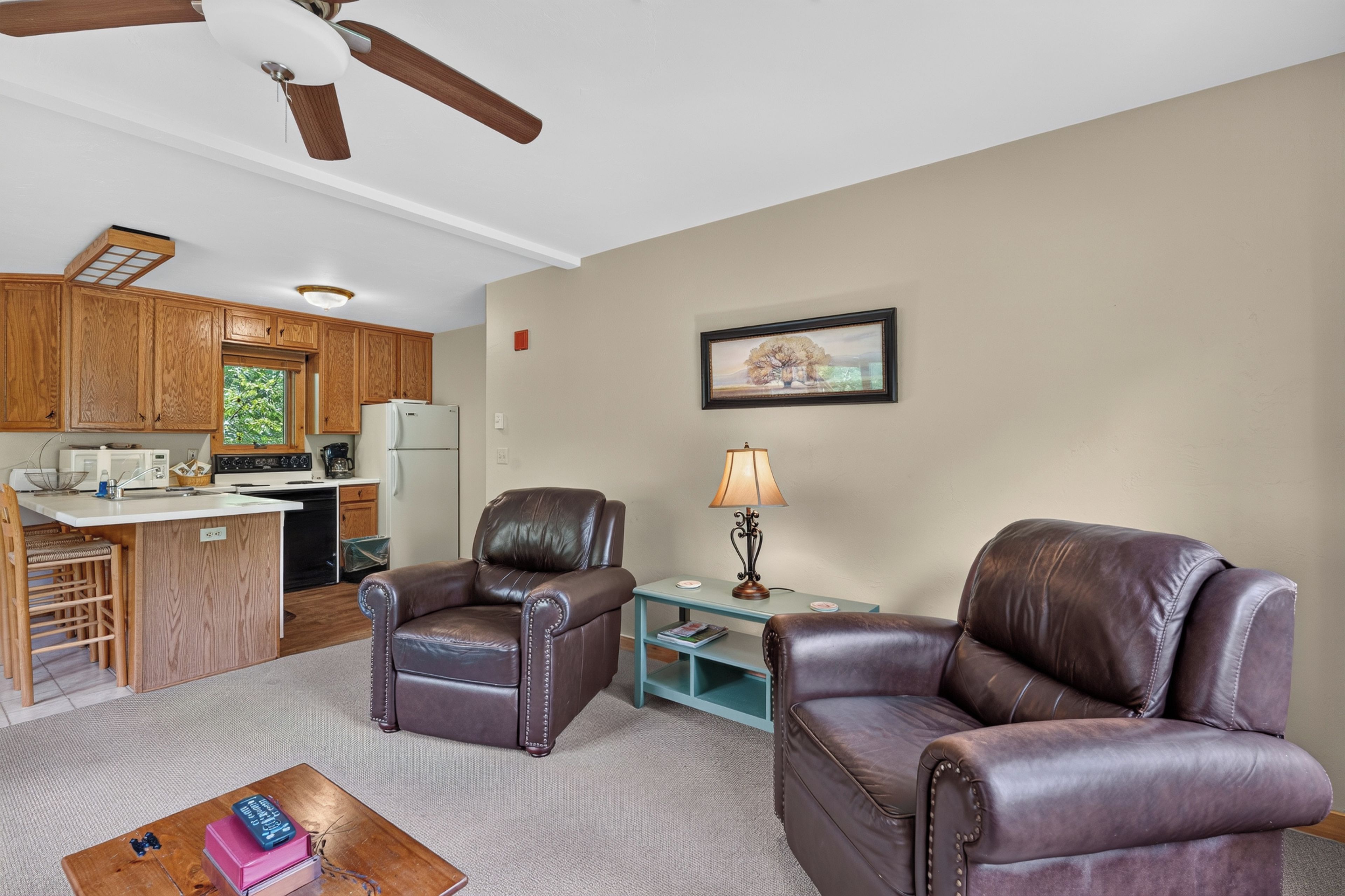 A compact living space with a kitchenette, two leather armchairs, a coffee table, and a framed picture on the wall.