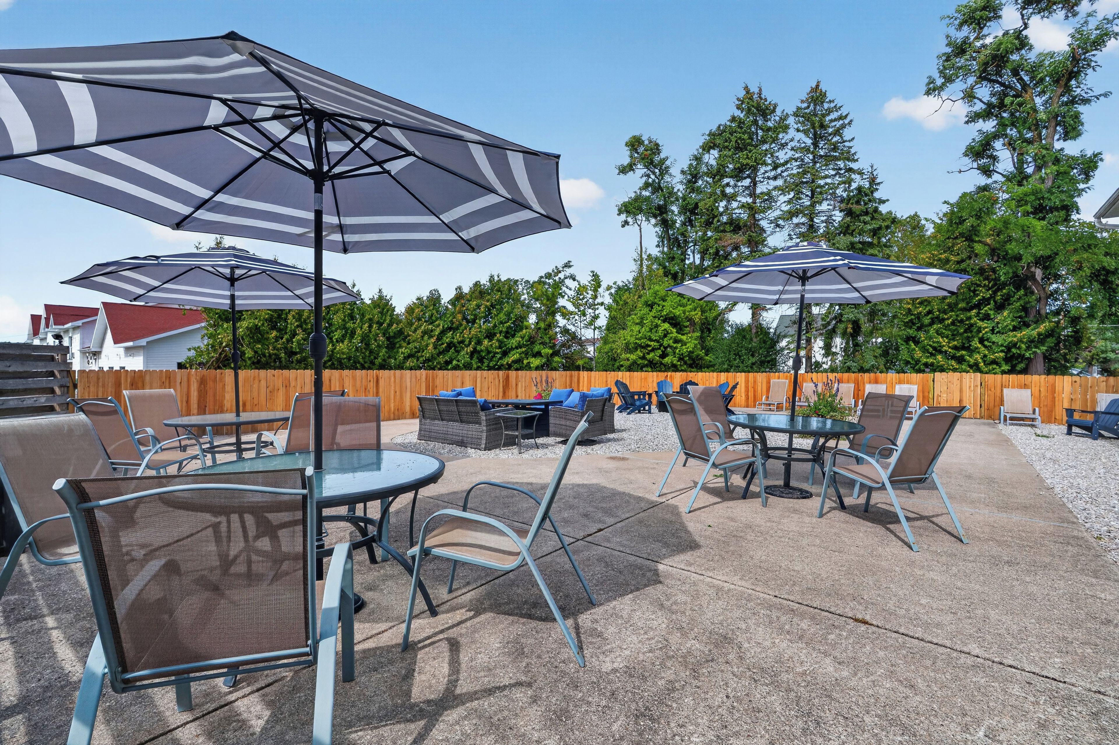 An outdoor patio area with round tables and chairs, large striped umbrellas, and a wooden fence in the background.