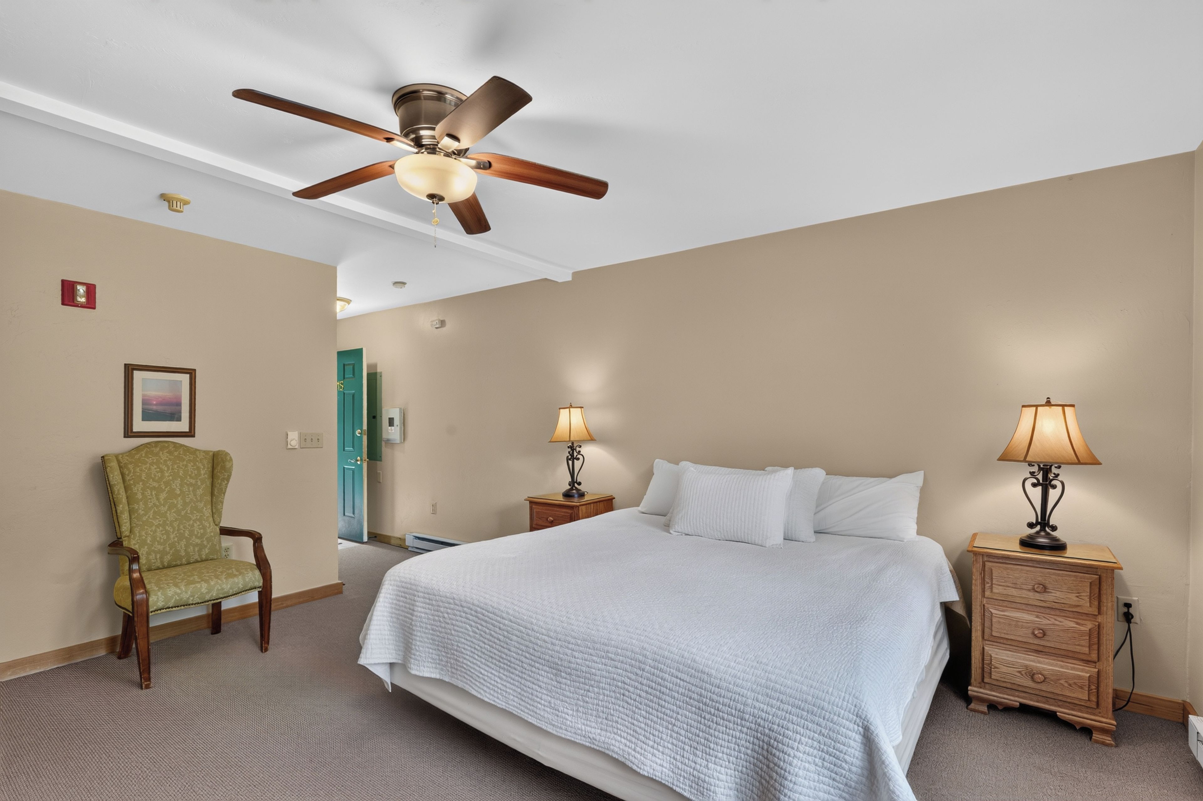 Habitación de hotel con cama, dos mesitas de noche con lámparas, una silla, ventilador de techo y una puerta.
