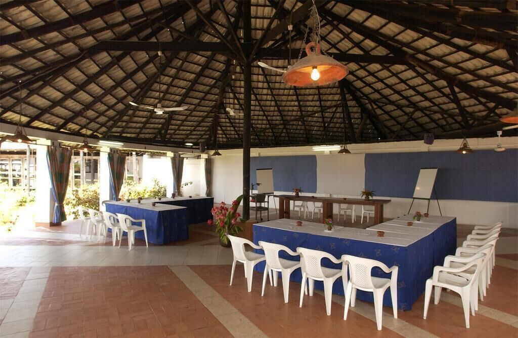 A covered outdoor dining area with a high ceiling, white plastic chairs, and a central table with a blue tablecloth.