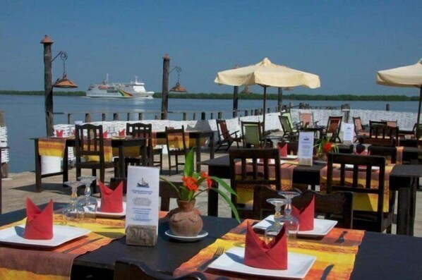 An outdoor dining area with tables set for two, umbrellas, and a view of the water with a boat in the background.
