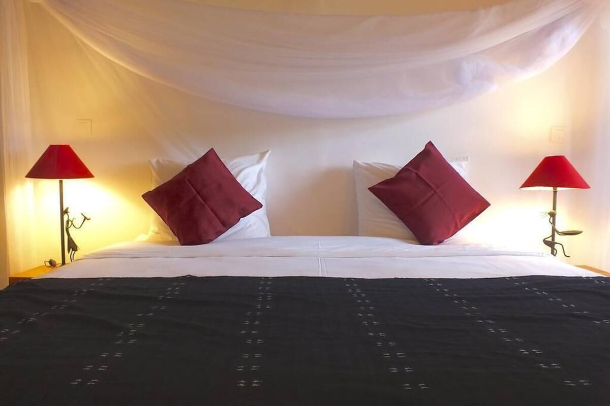 Un lit avec du linge de lit blanc et des oreillers rouges, deux lampes rouges de chaque côté, et une couvre-lit noire à motifs.