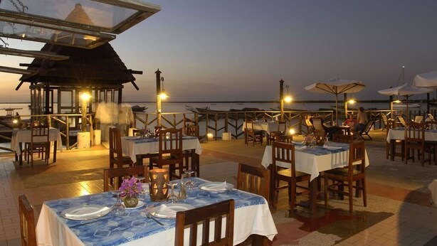 Un área de comedor al aire libre con mesas y sillas, una estructura de techo de paja y vista al mar.