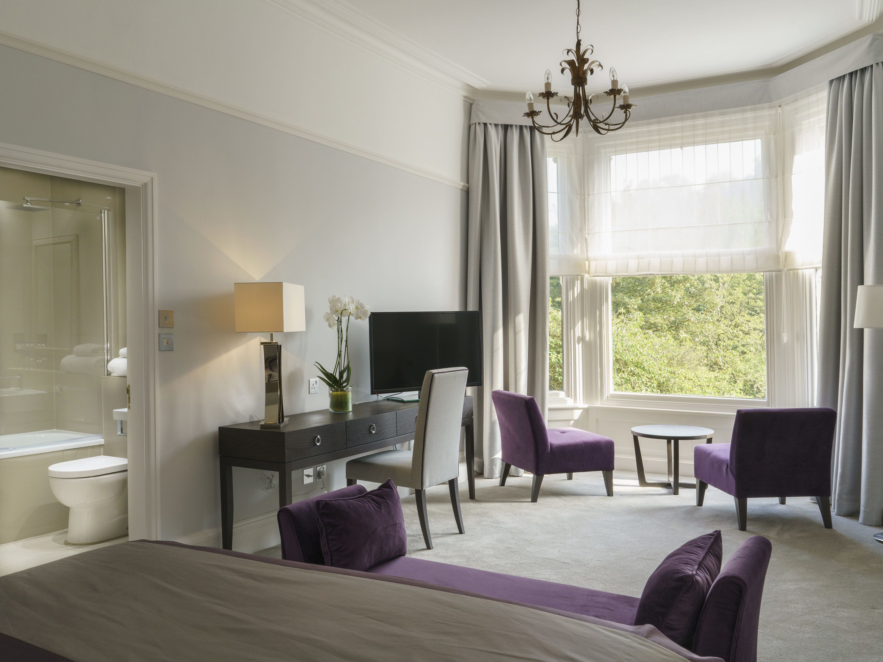 A hotel room with a bed, a desk with a TV, a chair, a small table, and a window with curtains.