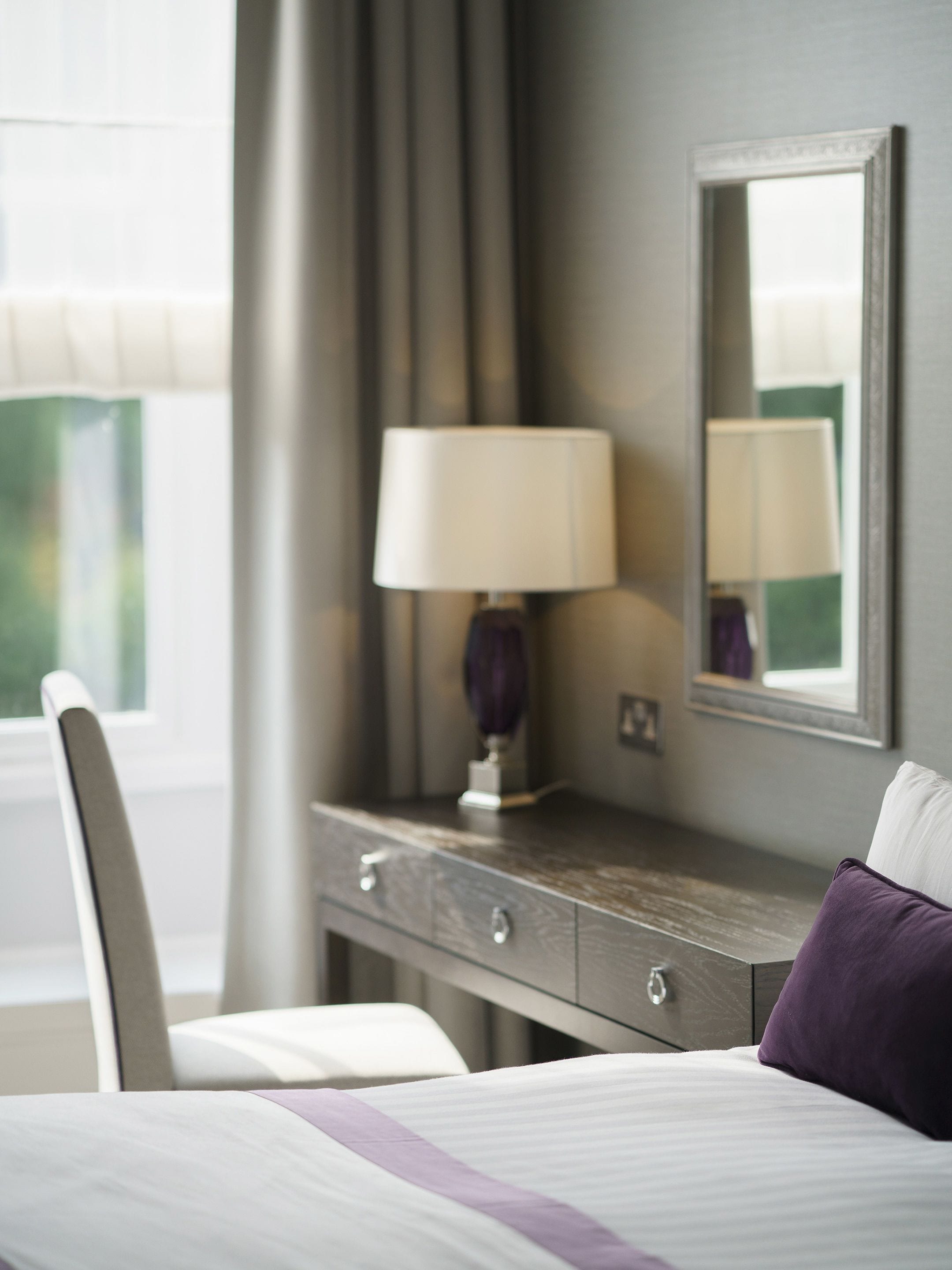 A hotel room with a bed, a bedside table with a lamp, a chair, and a mirror on the wall.