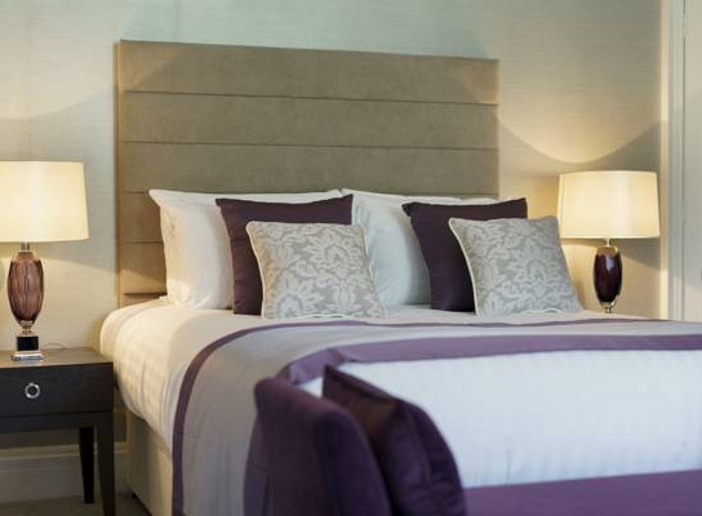 A neatly made bed with a headboard, two bedside tables with lamps, and a purple throw blanket.