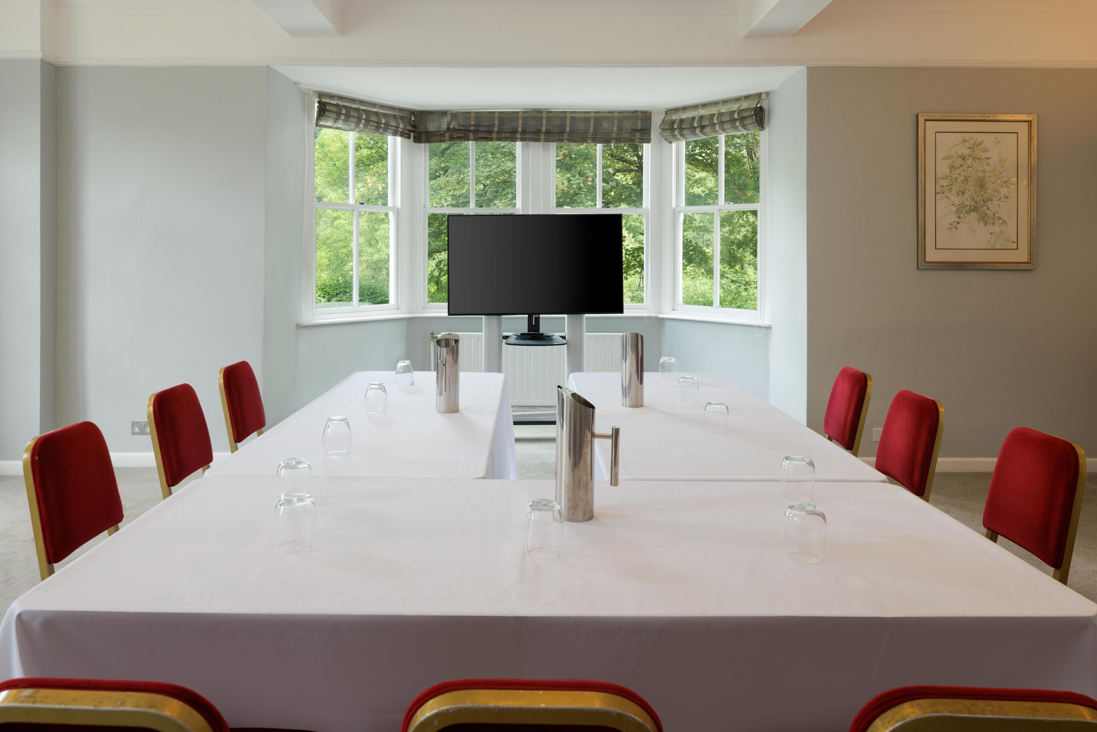 A conference room with a long table, red chairs, a flat-screen TV, and large windows with a view of greenery.