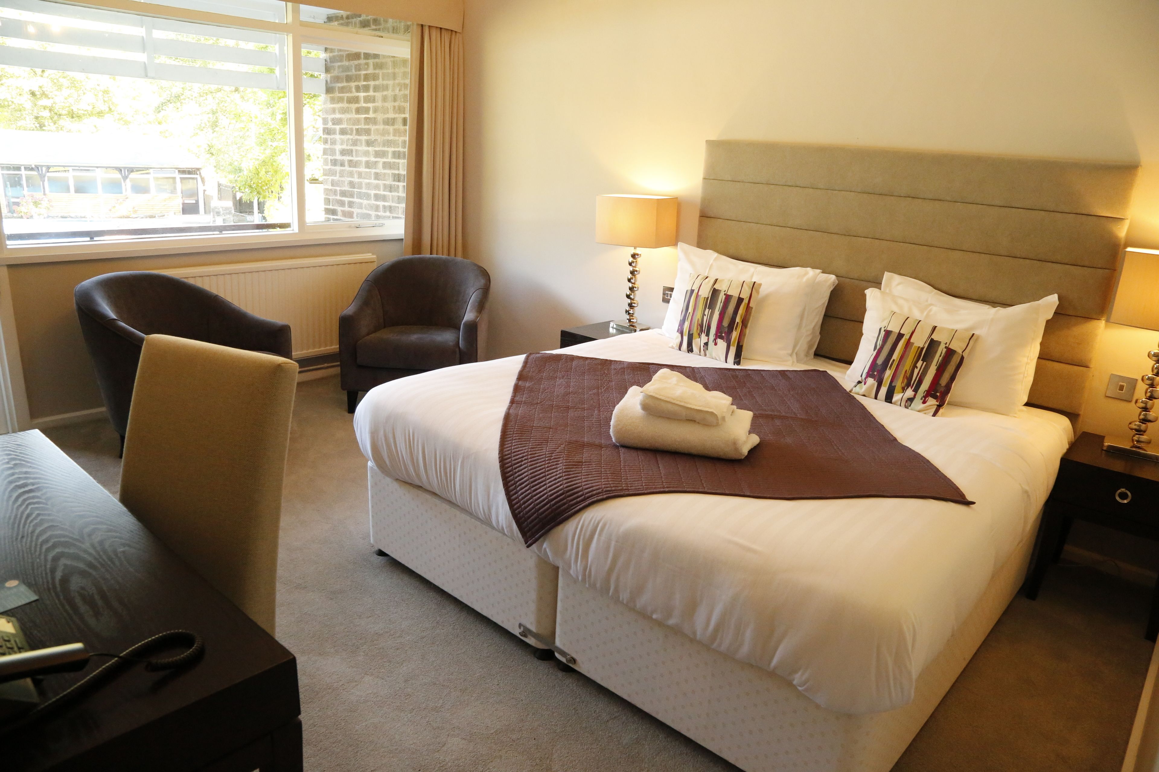 A hotel room with a large bed, a chair, a desk with a telephone, and a window with curtains.