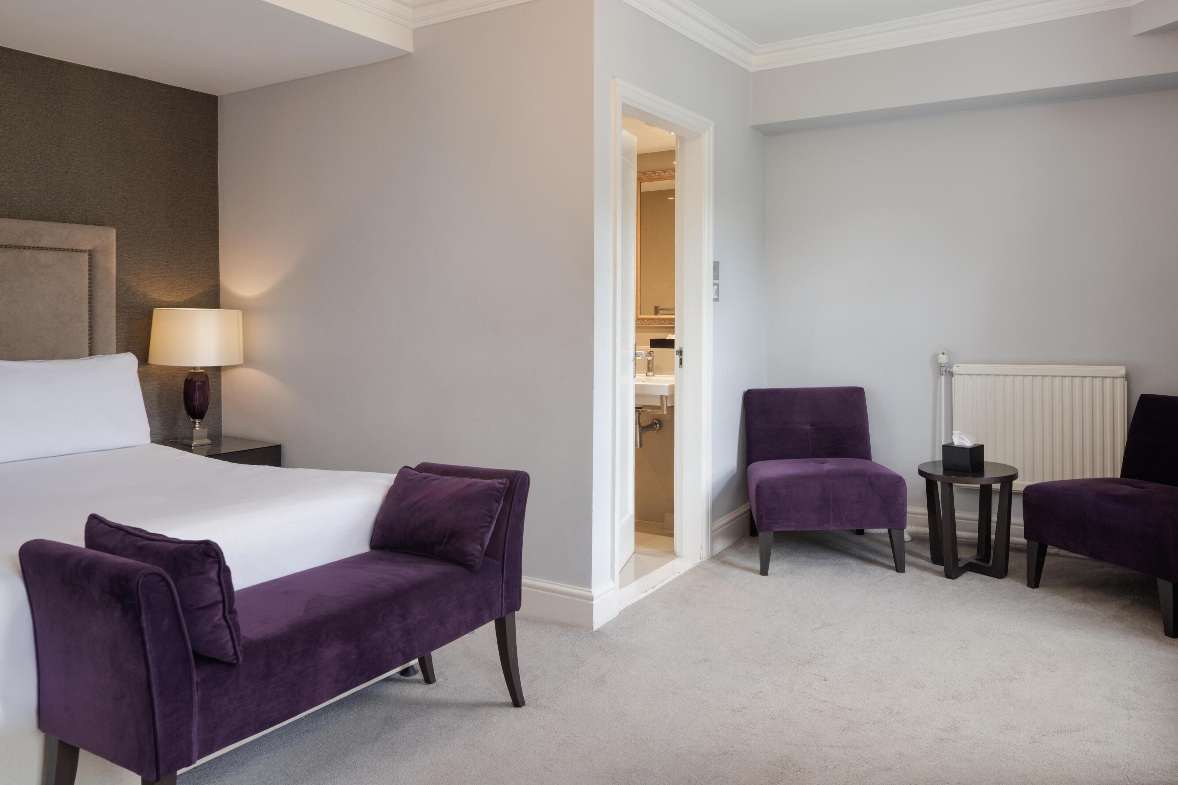 A hotel room with a bed, two purple armchairs, a small table with a lamp, and a radiator.