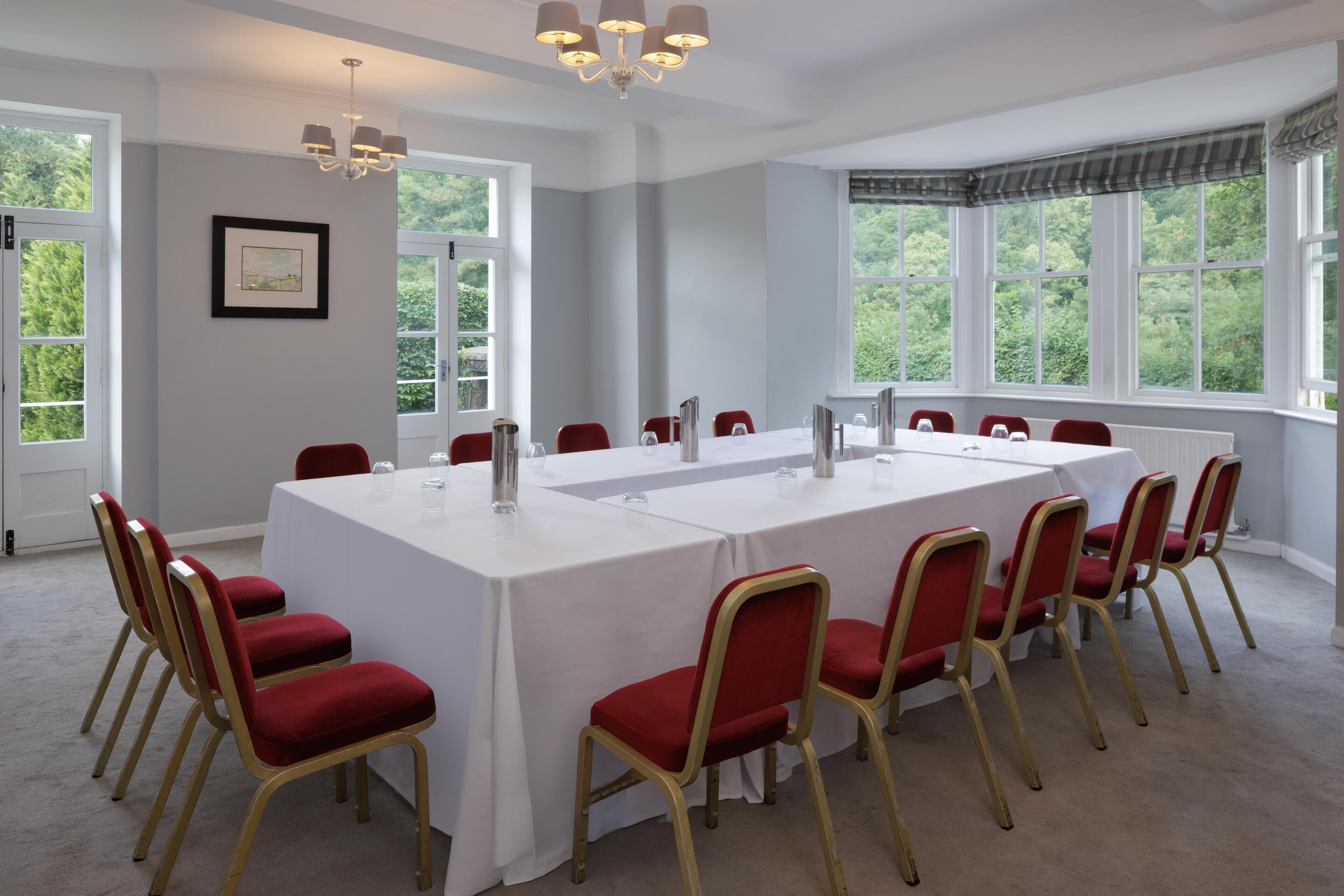 A conference room with a long table, red chairs, and large windows with a view of greenery.