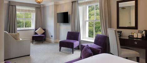 A hotel room with a large bed, two purple armchairs, a grey sofa, a desk with a mirror, and a TV mounted on the wall.