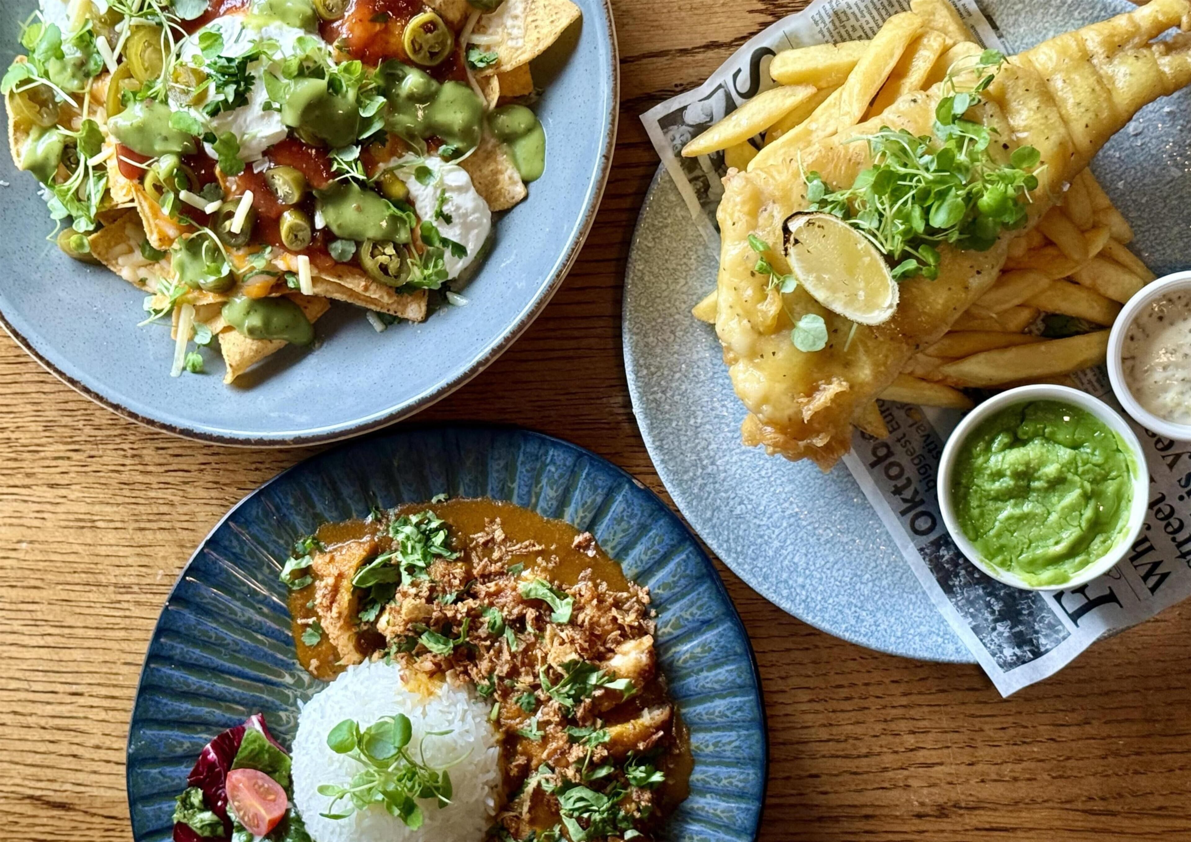 A table with three plates of food: nachos, fish and chips, and a plate with rice and a meat dish.