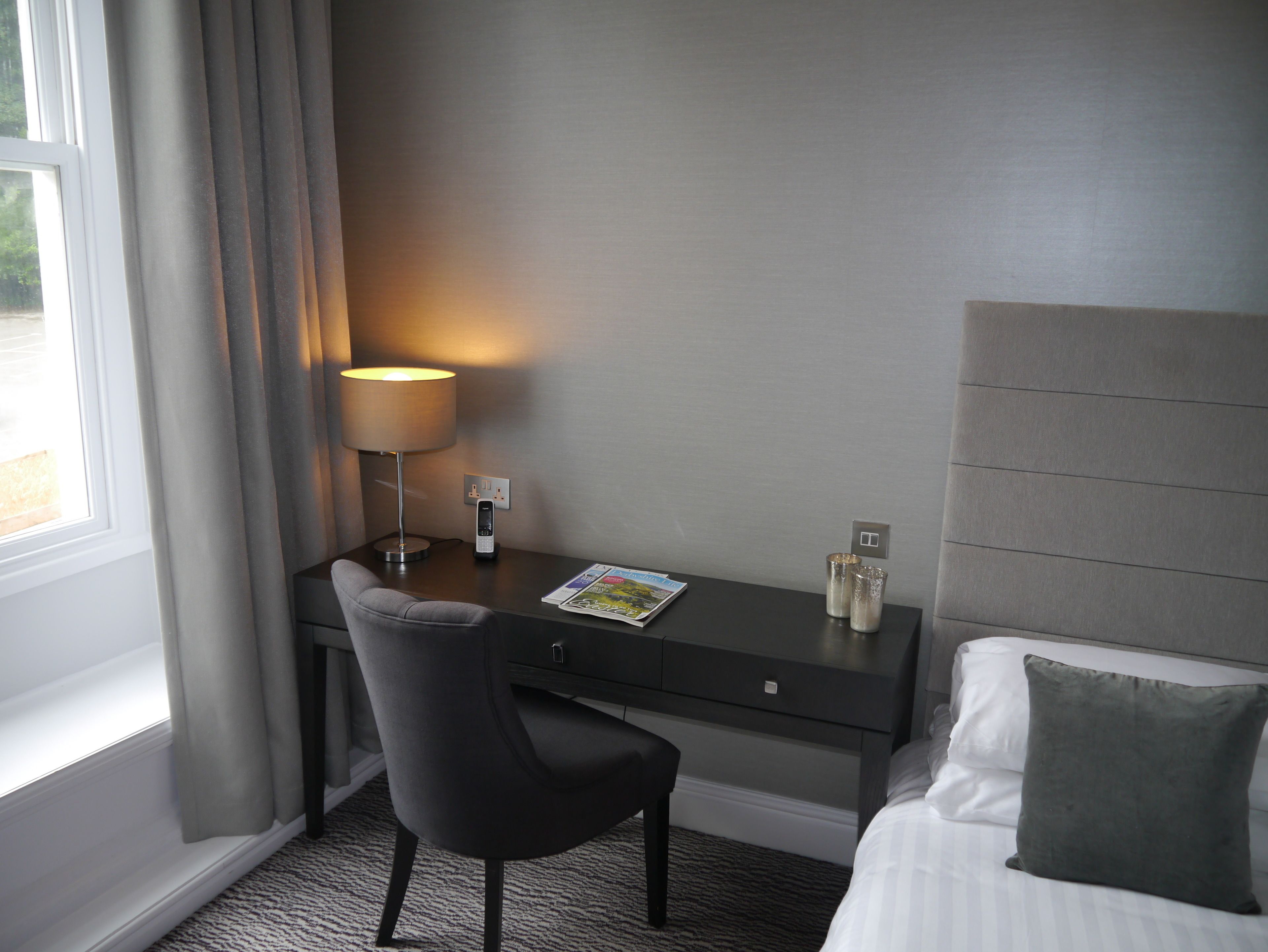 A hotel room with a desk, chair, lamp, and a bed with pillows.
