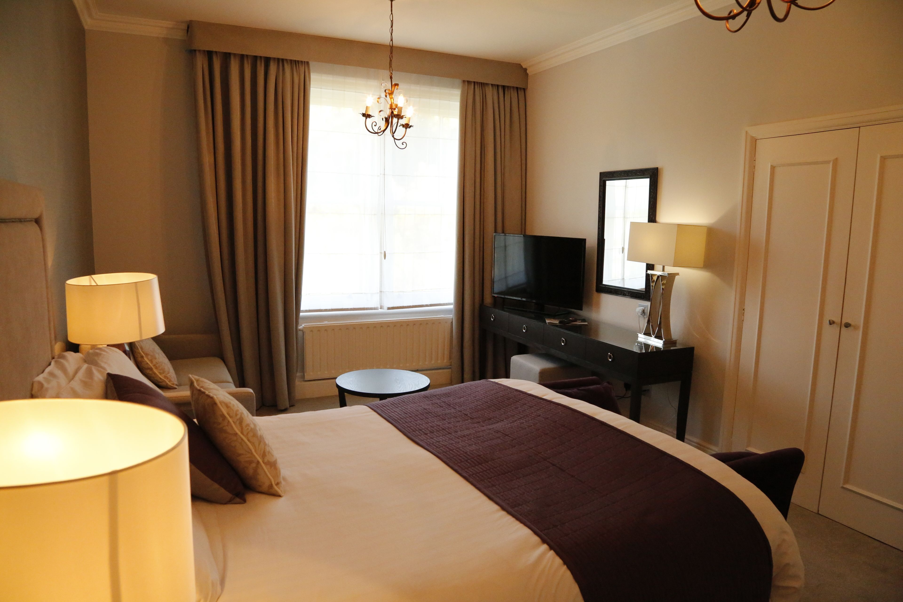 A hotel room with a large bed, a desk with a TV, a chair, a lamp, and a window with curtains.