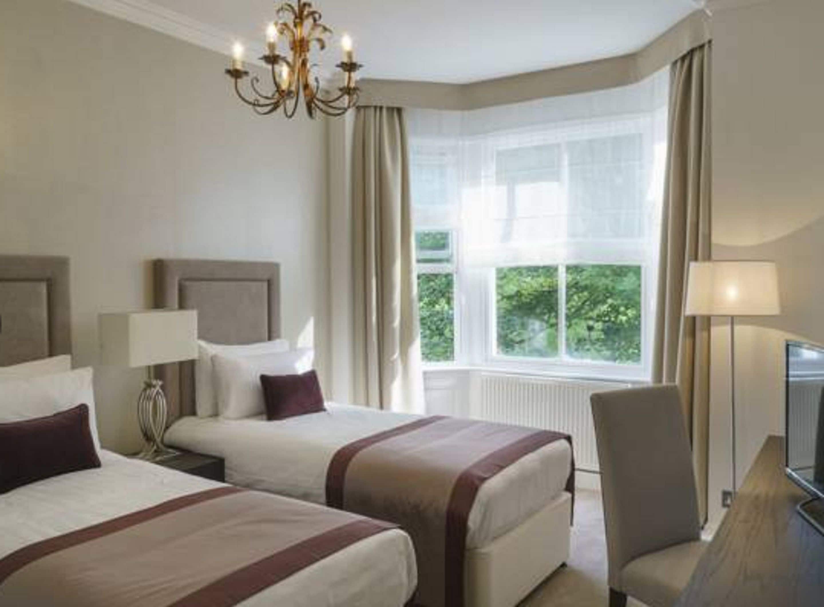 Habitación de hotel con dos camas, un candelabro, un ventanal grande con cortinas y un televisor.