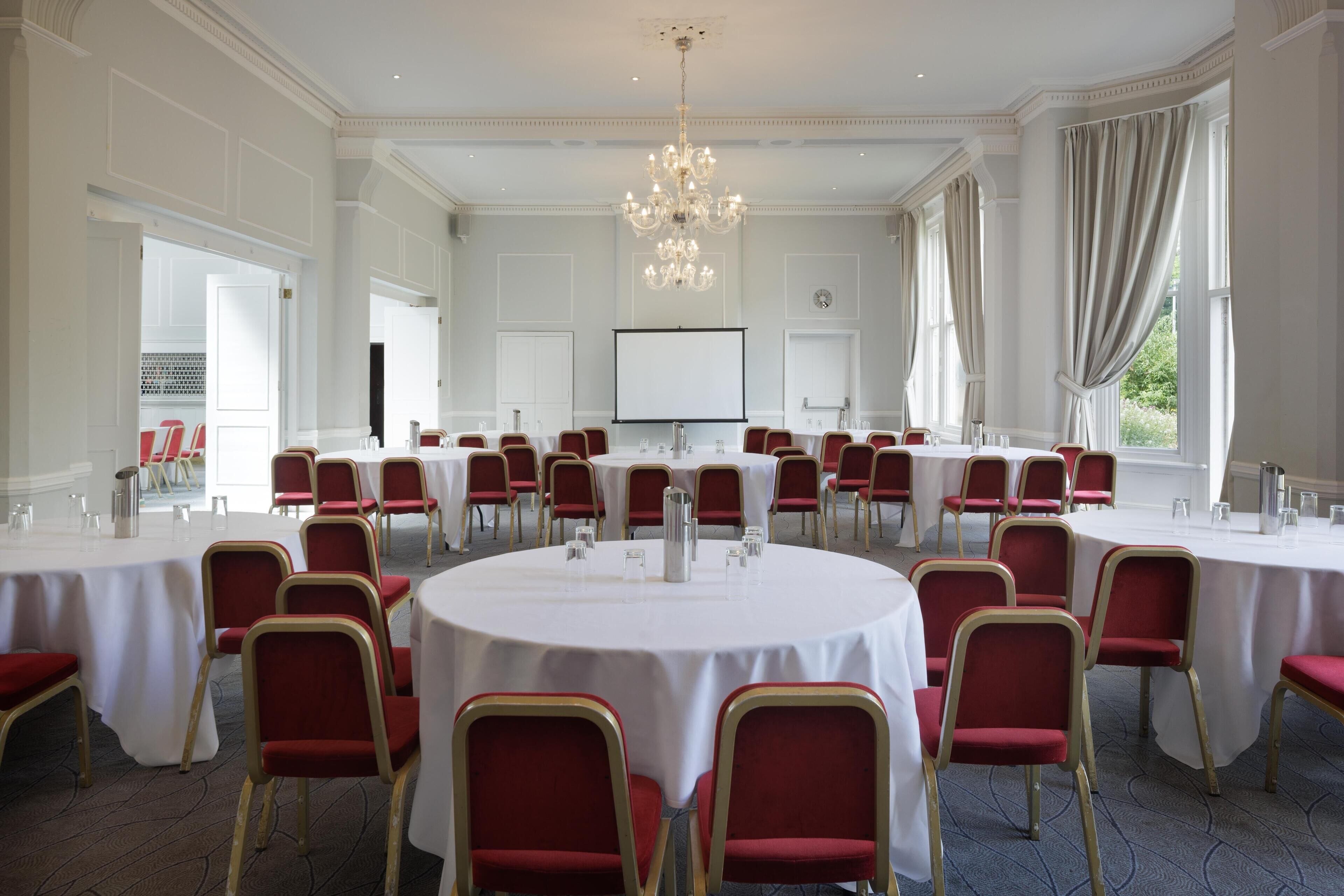 A large conference room with round tables and red chairs, a chandelier, and a projector screen.