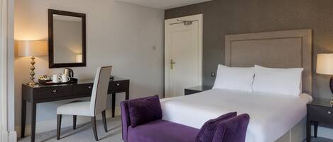 A hotel room with a bed, a desk, a chair, a purple bench, a nightstand, a lamp, and a door.