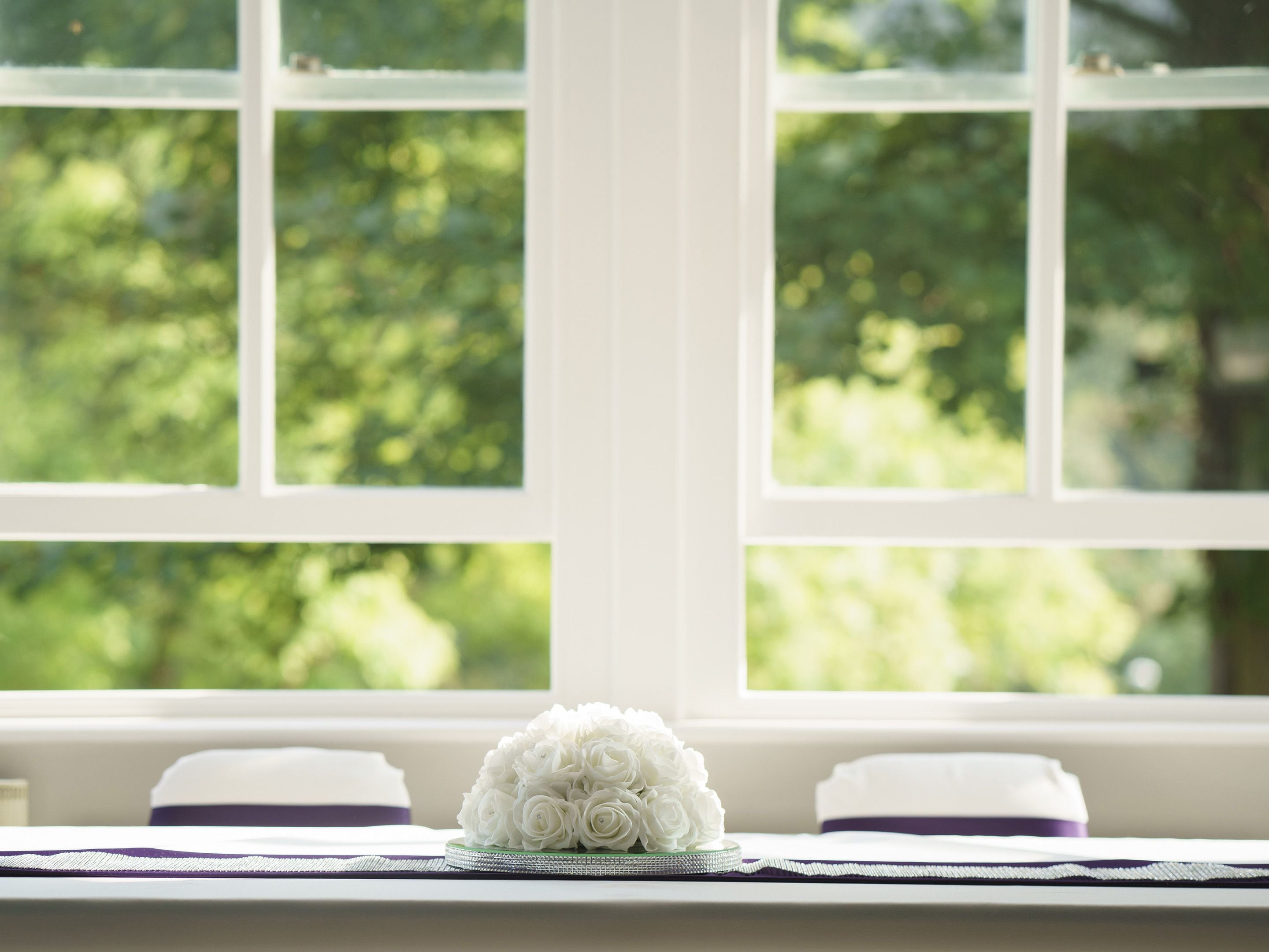 A window with a view of greenery, a white table with a glass vase of white flowers, and a purple and white pillow.