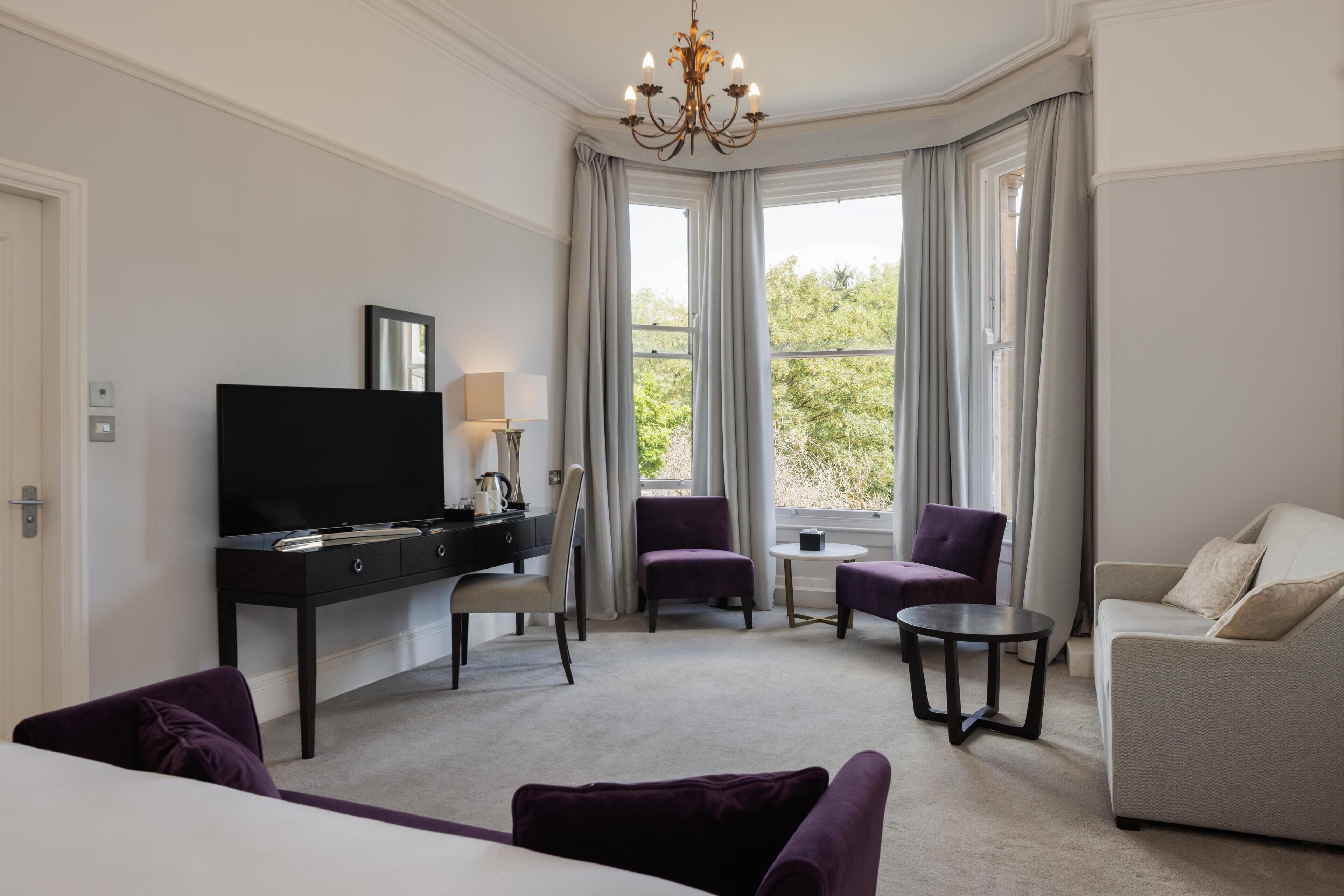 A modern hotel room with a flat-screen TV, a desk, a sofa, a chair, and a small round table.