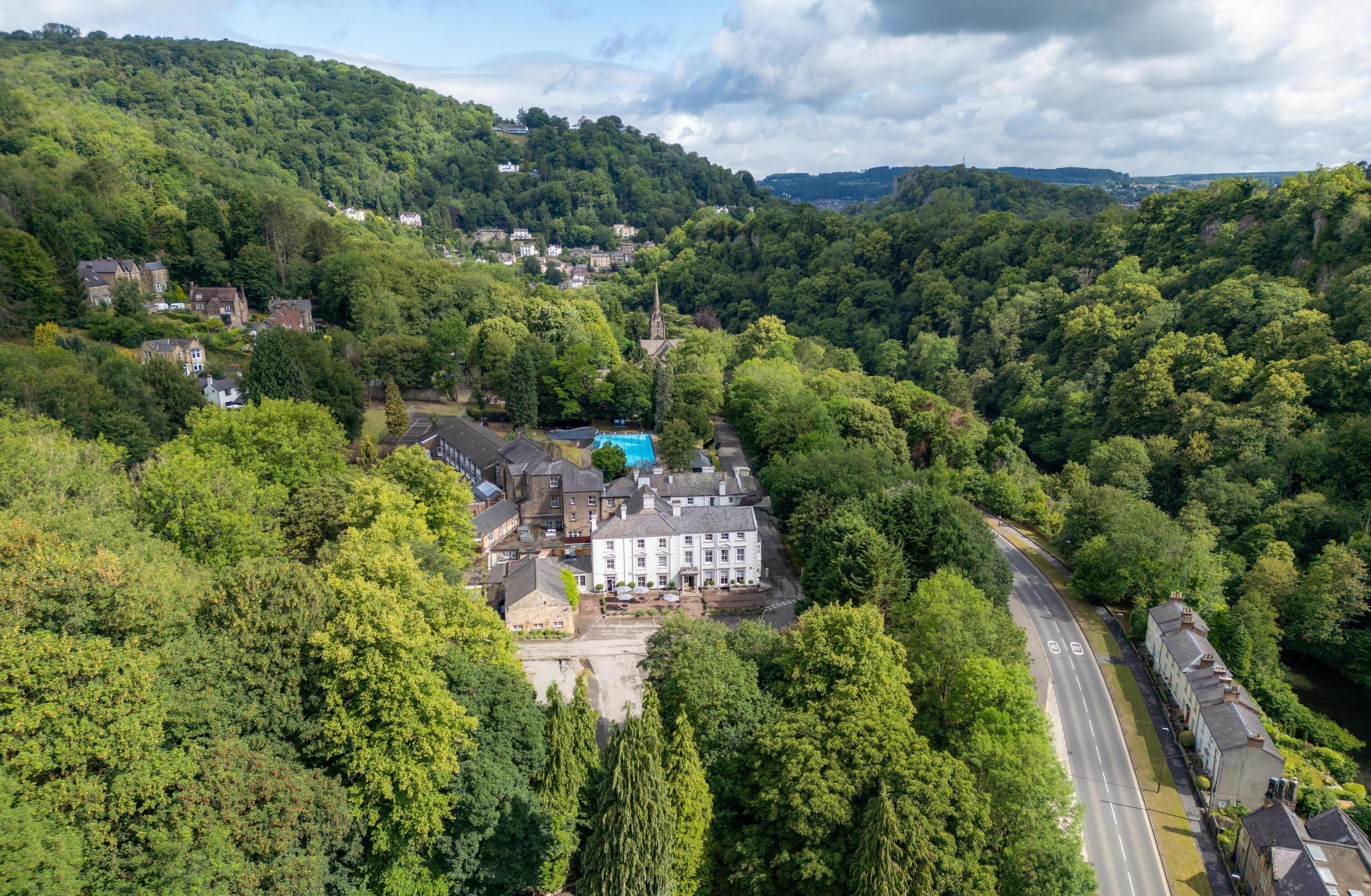 A large, white building with a swimming pool, surrounded by dense greenery and a road.
