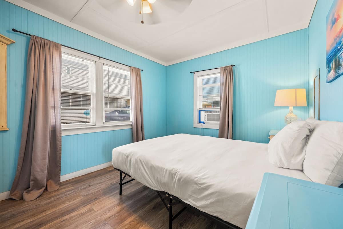 A bedroom with a bed, two windows with curtains, a ceiling fan, and a wall-mounted lamp.