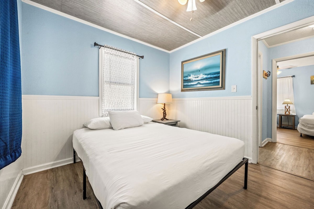 A bedroom with a large bed, a window with blinds, a painting on the wall, and a lamp on a bedside table.