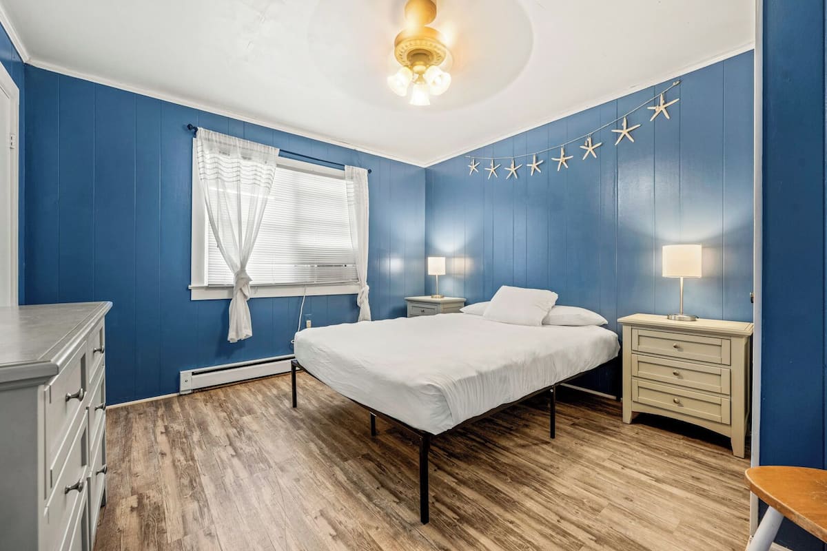 A bedroom with a bed, bedside tables, a nightstand, a window with curtains, and a ceiling fan.