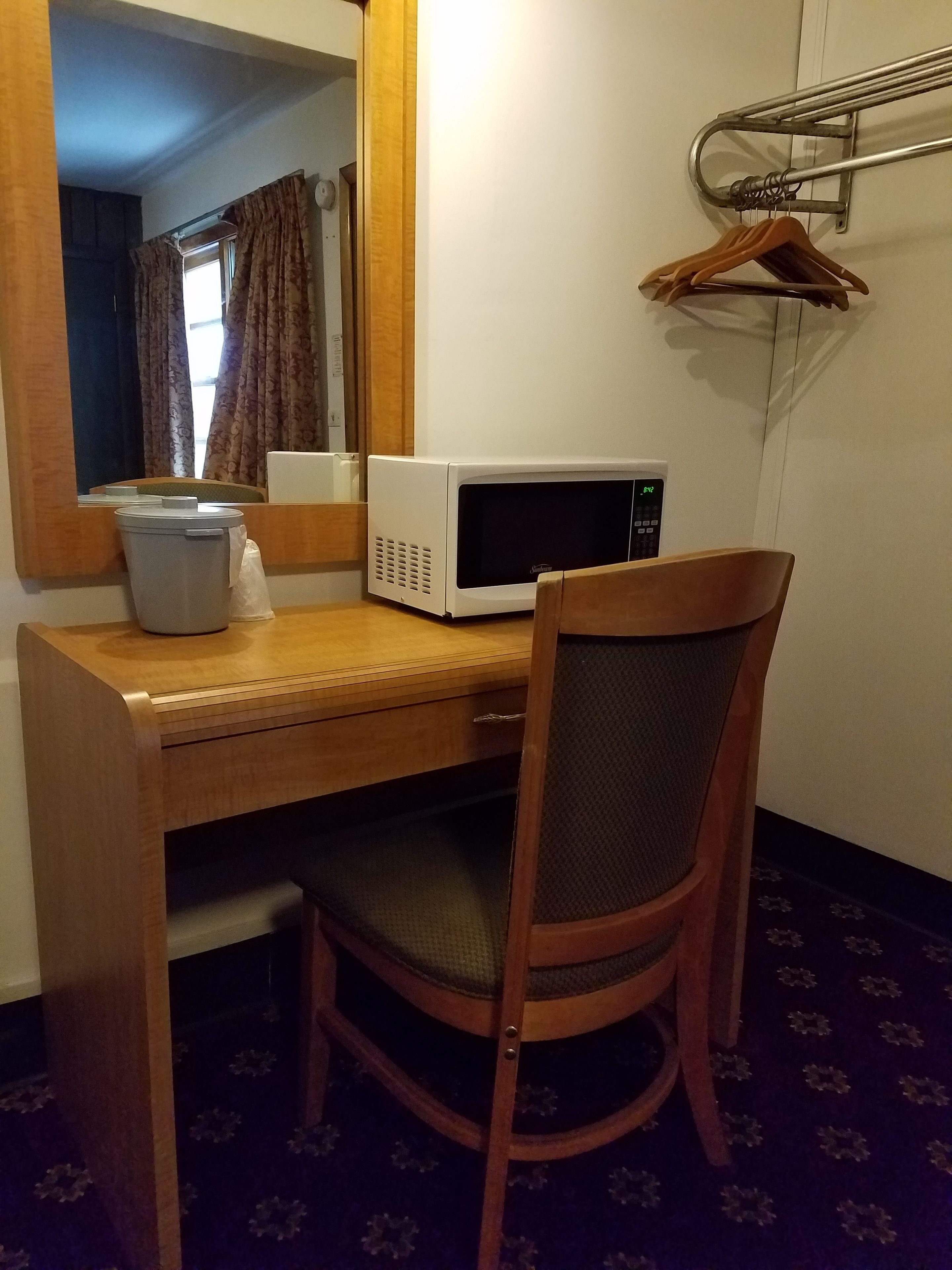 Et lille hotelværelse med skrivebord i træ, mikroovn, stol, tøjstativ og skraldespand.