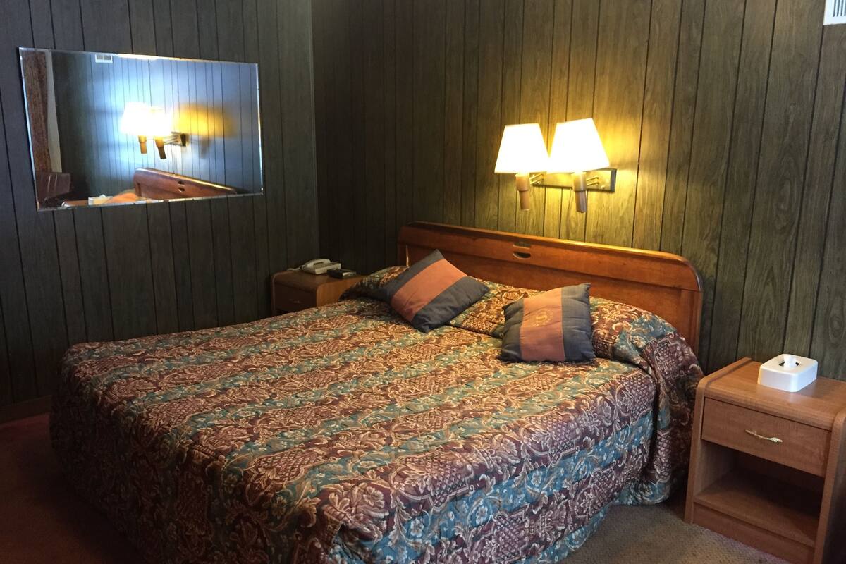 Habitación de hotel con una cama grande, cabecera de madera, dos lámparas de noche, una mesita de noche con teléfono y lámpara, y televisor montado en la pared.