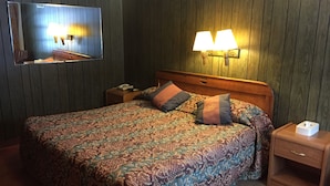 Habitación de hotel con una cama grande, cabecera de madera, dos lámparas de noche, una mesita de noche con teléfono y lámpara, y televisor montado en la pared.