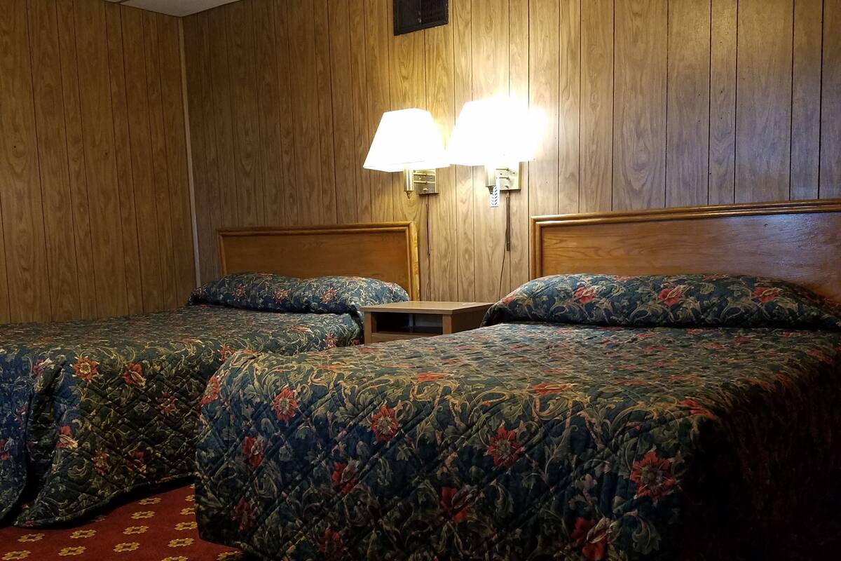 Habitación de hotel con dos camas, paneles de madera, alfombra roja con estampado floral y dos lámparas fijadas a la pared.