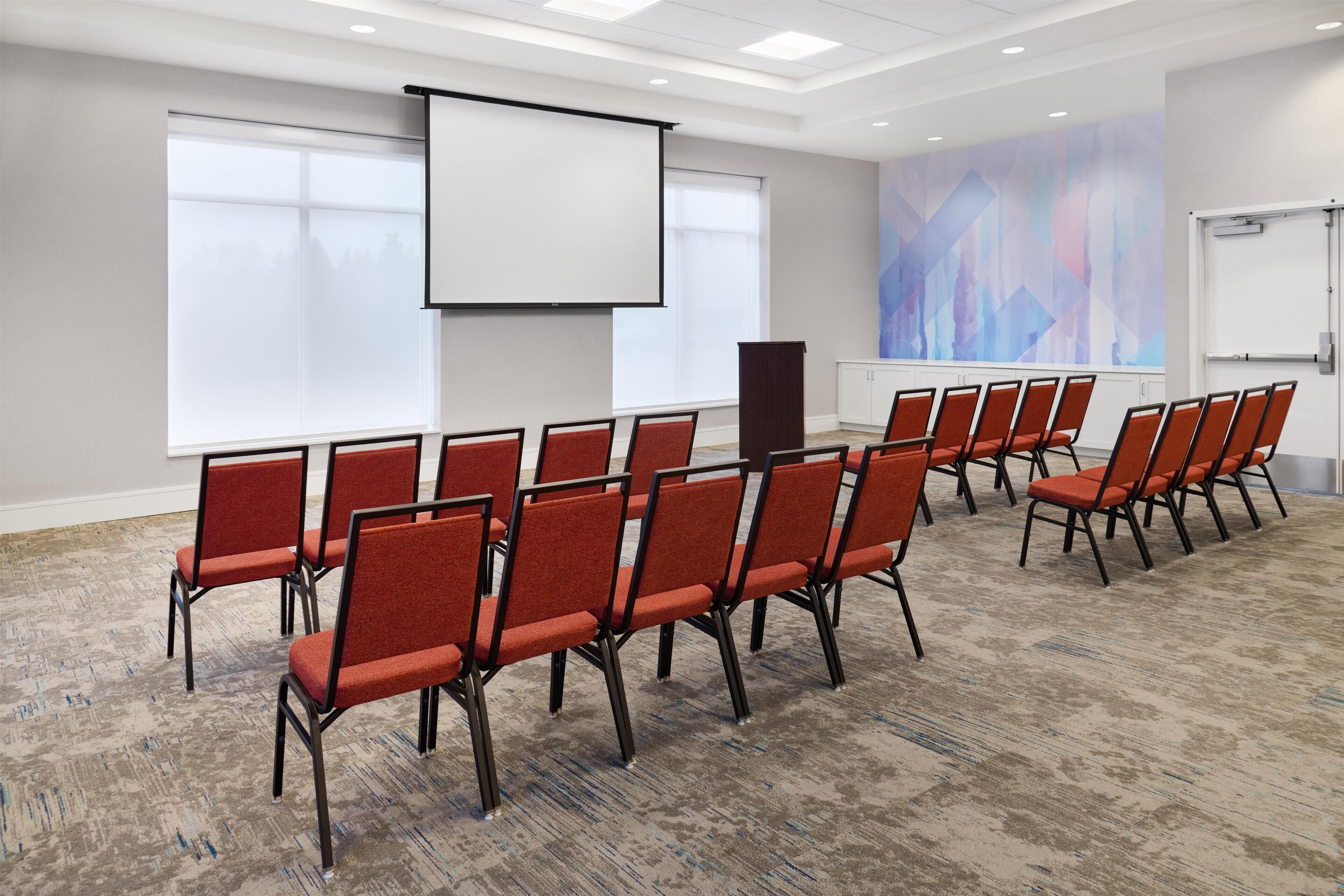 A conference room with rows of chairs, a projector screen, a podium, and large windows.