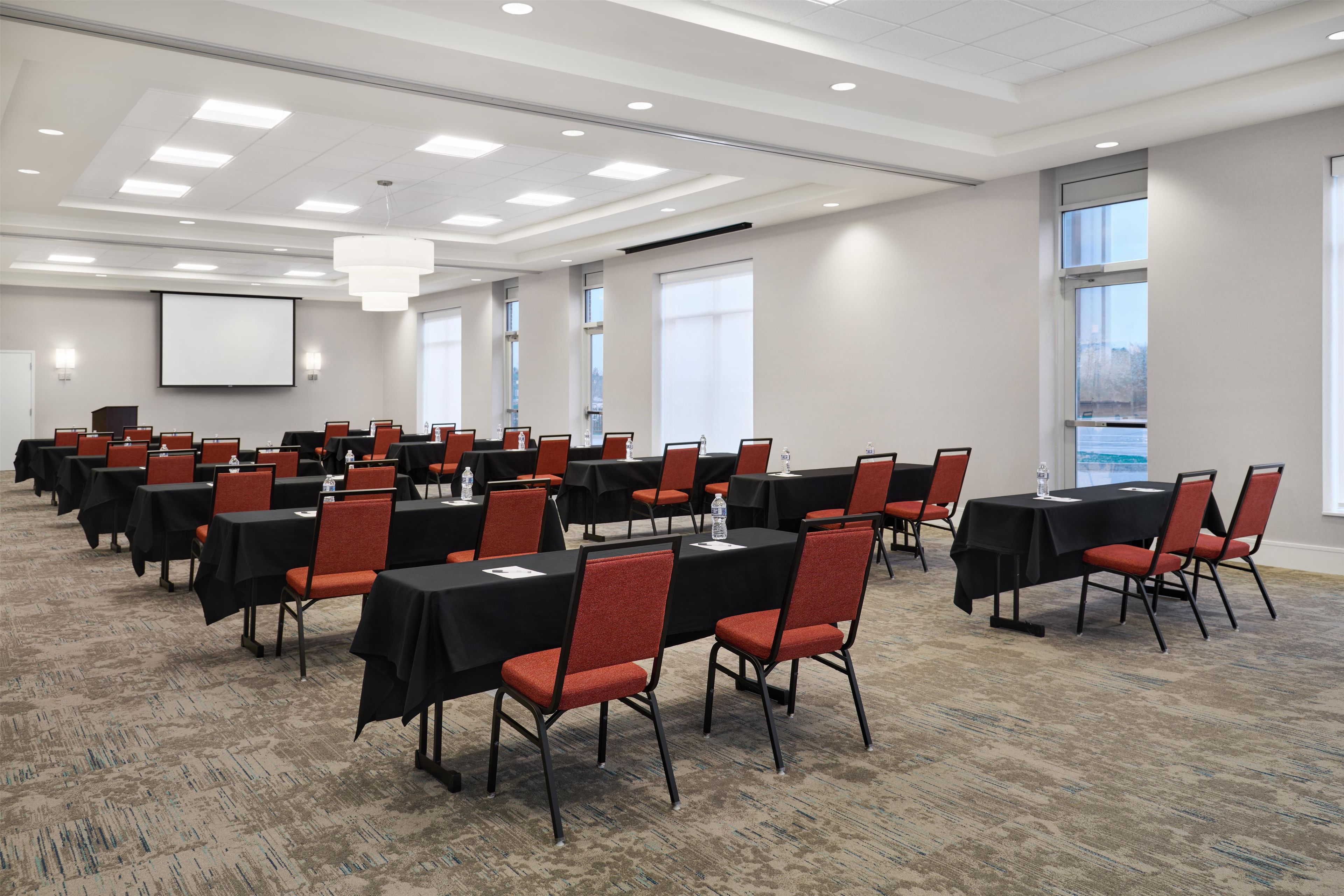 A conference room with multiple tables and chairs, a projector screen, and large windows.