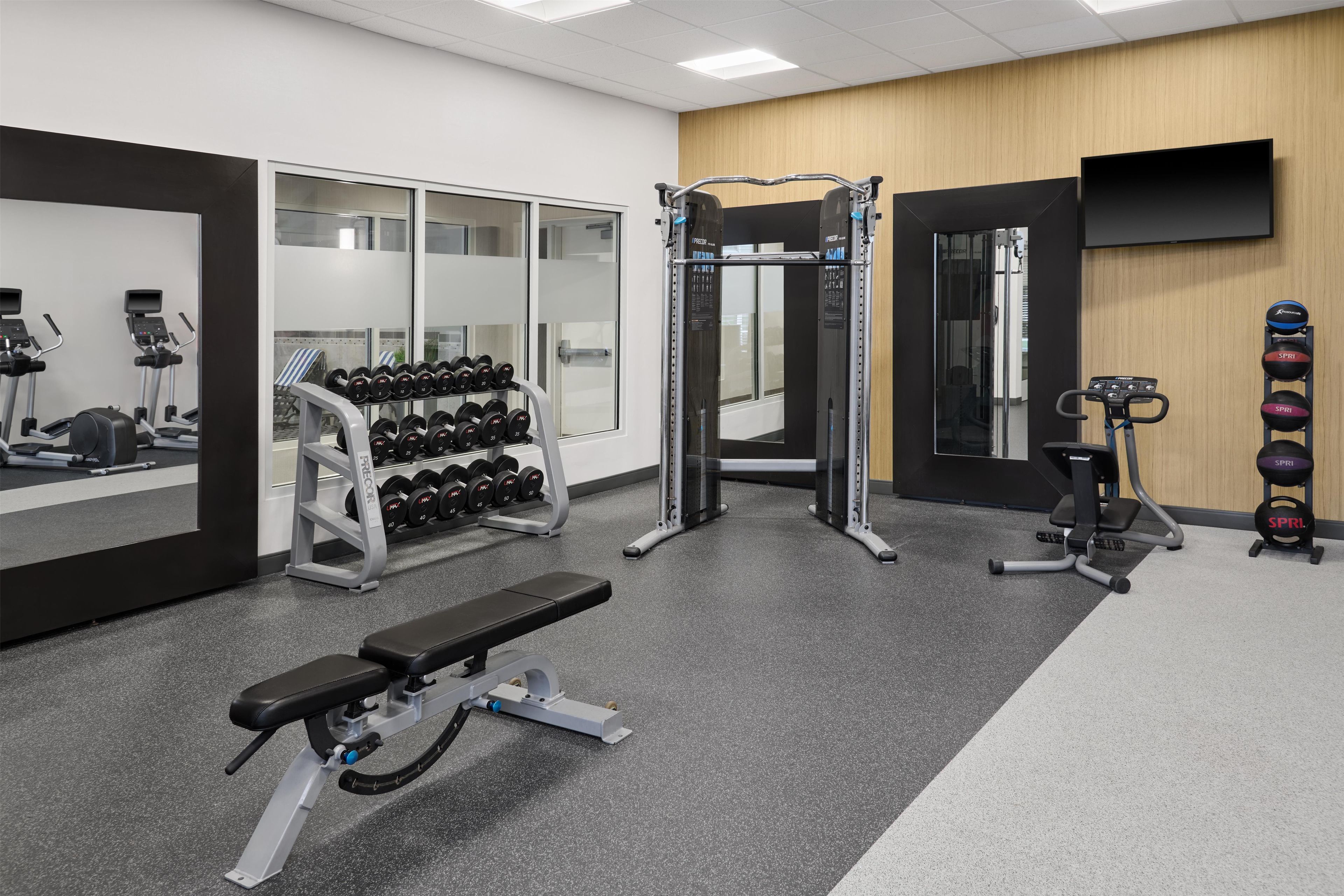 Un gimnasio con equipo de ejercicio, una banca, mancuernas y un televisor en la pared.