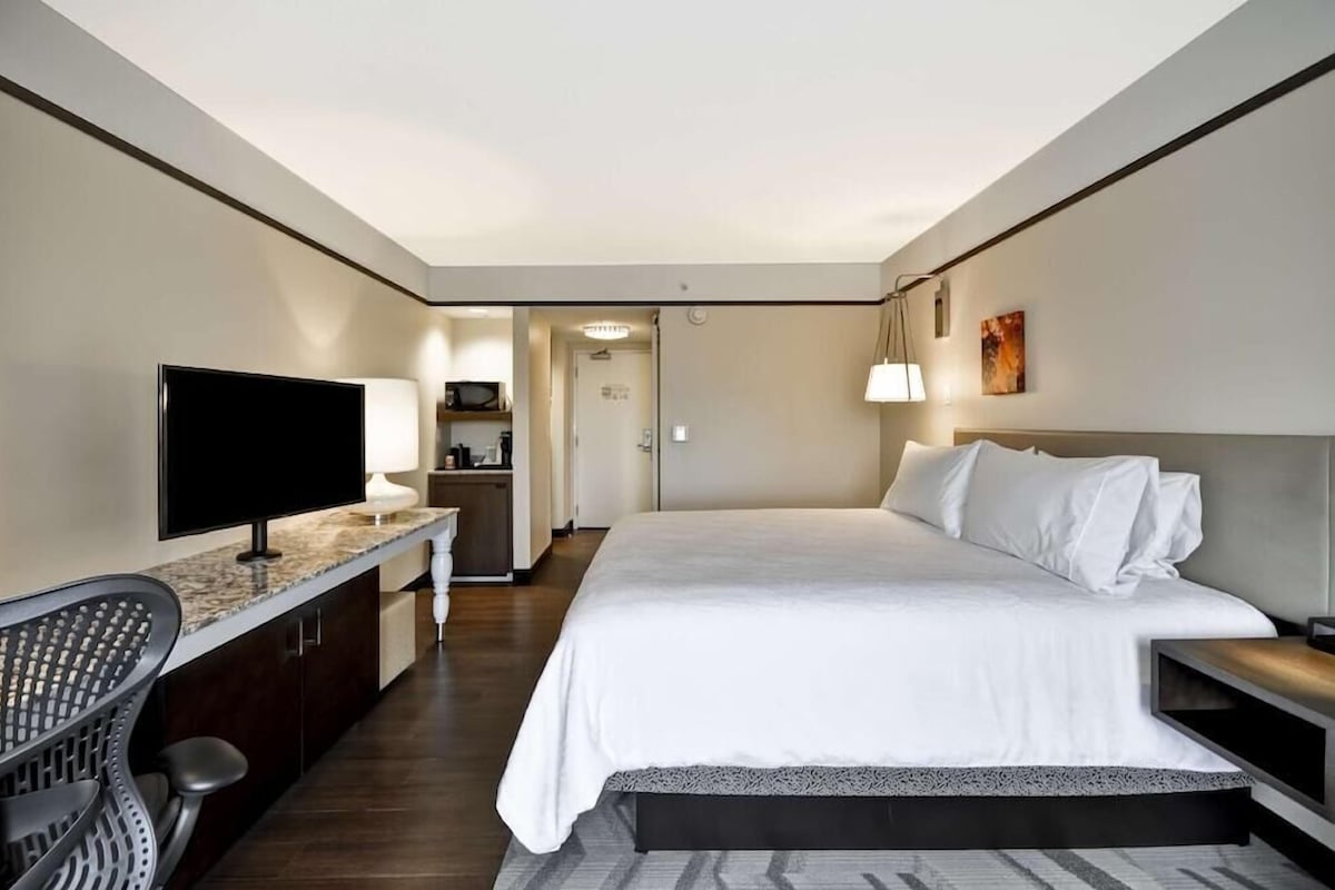 A hotel room with a bed, a TV, a desk with a chair, and a kitchenette in the background.