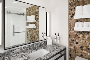 A modern bathroom with a granite countertop, a glass shower enclosure, and a stone wall with towel racks. - Hilton Garden Inn Detroit Troy (Troy)