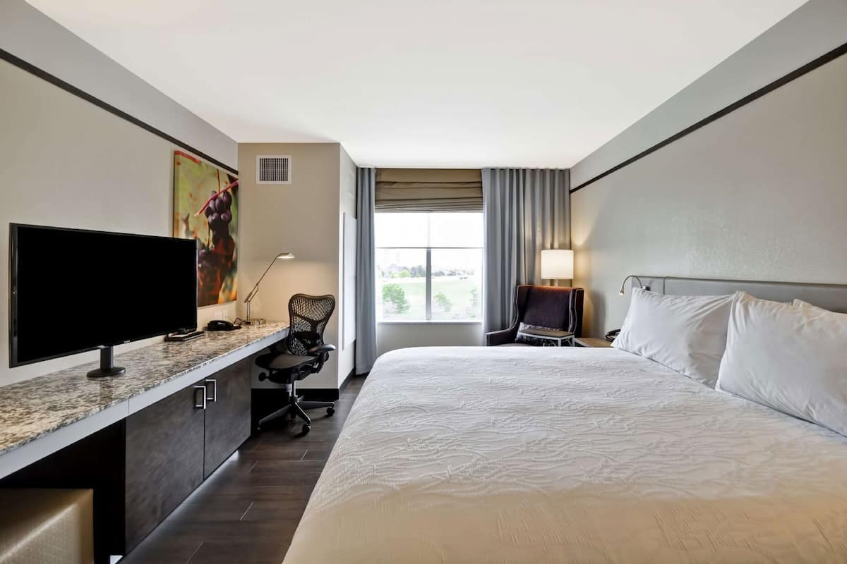 A hotel room with a large bed, a desk with a flat-screen TV, a chair, a lamp, and a window with curtains.