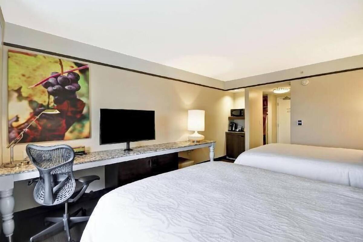 A hotel room with a bed, desk, chair, TV, and a painting of grapes.