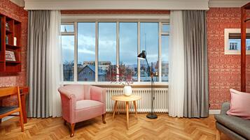 A room with a large window, a wooden floor, a red patterned wall, a white radiator, a round table, a lamp, a red and white chair, and a grey sofa.