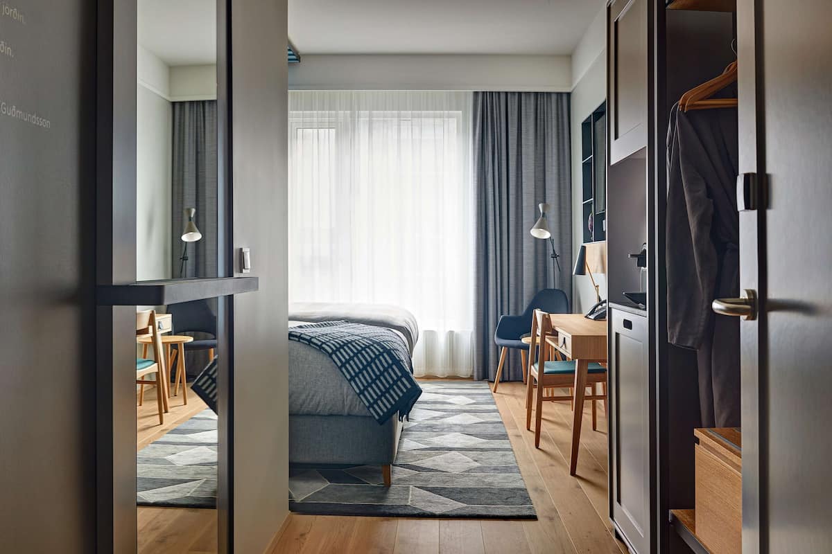 Een moderne hotelkamer met een bed, bureau en stoel. Er is een groot raam met gordijnen, een spiegel en een kledingkast.